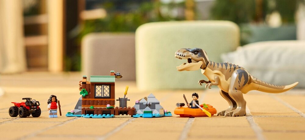 Zestaw LEGO przedstawiający scenę z tyranozaurem, łodzią, małym budynkiem i quadem. W tle widoczne są rozmyte elementy otoczenia.