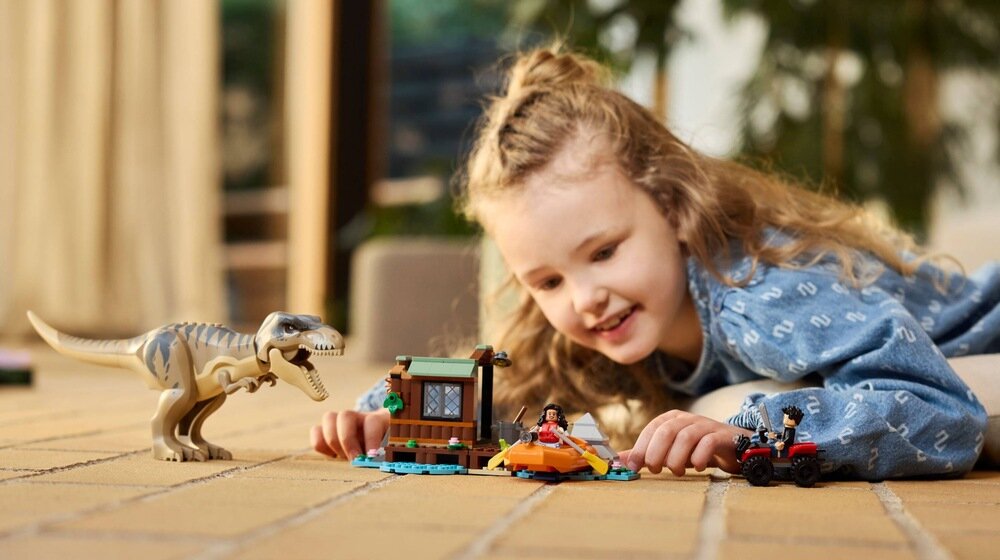Dziecko bawi się zestawem LEGO, który zawiera figurkę tyranozaura, łódź i mały domek. W tle widać roślinność domową.