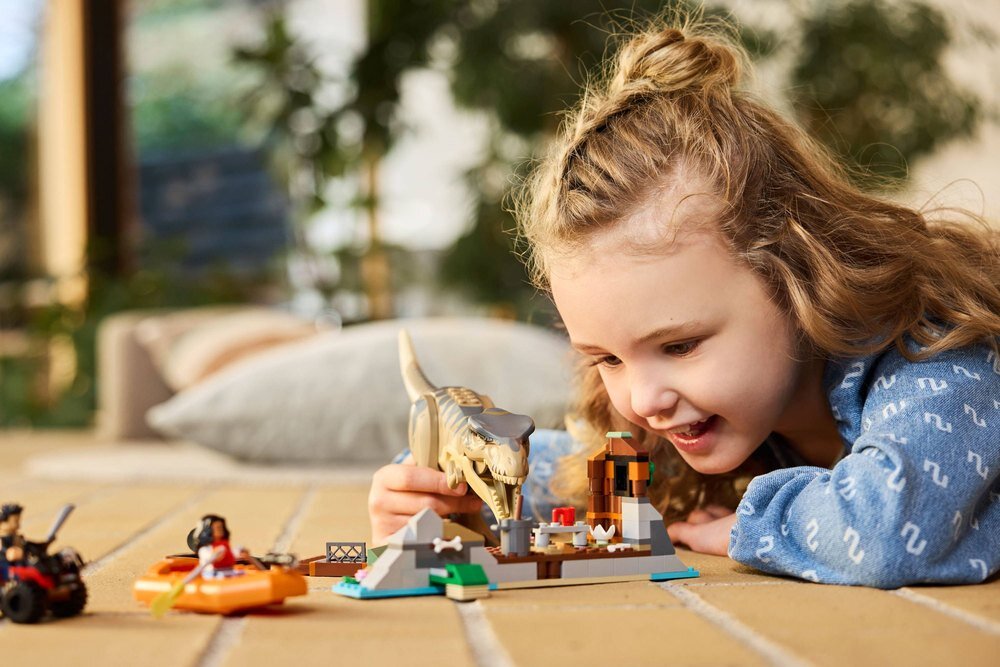 Dziecko bawi się zestawem klocków LEGO z motywem dinozaura i małej łodzi. W tle widoczne są rozmyte elementy wnętrza.