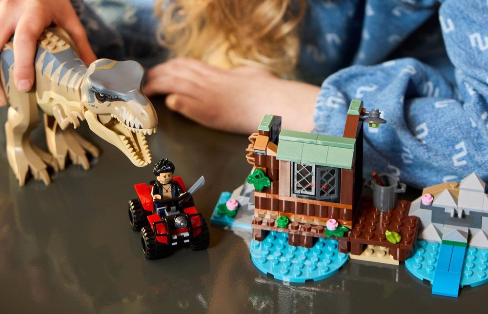 Na obrazie widać zestaw klocków LEGO przedstawiający tyranozaura, figurkę na quadzie oraz małą konstrukcję przypominającą domek nad wodą. W tle widoczne są ręce osoby w niebieskim ubraniu.