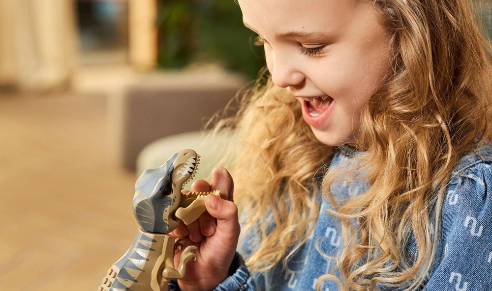 Dziecko trzyma figurkę dinozaura z klocków LEGO, uśmiechając się do niej. W tle widoczne są rozmyte elementy wnętrza.