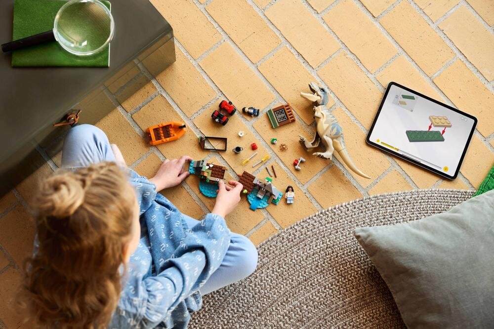 Dziecko bawi się zestawem klocków LEGO, w tym figurką tyranozaura i małymi pojazdami, na podłodze z jasnych płytek. Obok leży tablet z wyświetlonym schematem budowy z klocków.