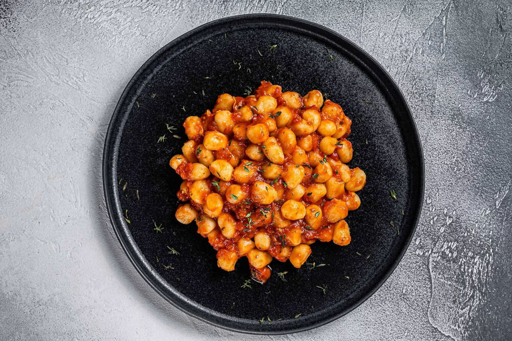 Plan dietetyczny BEDIET 1 miesiąc Włoskie kopytka gnocchi w gęstym sosie pomidorowym, posypane świeżymi ziołami, serwowane na minimalistycznym czarnym talerzu.
