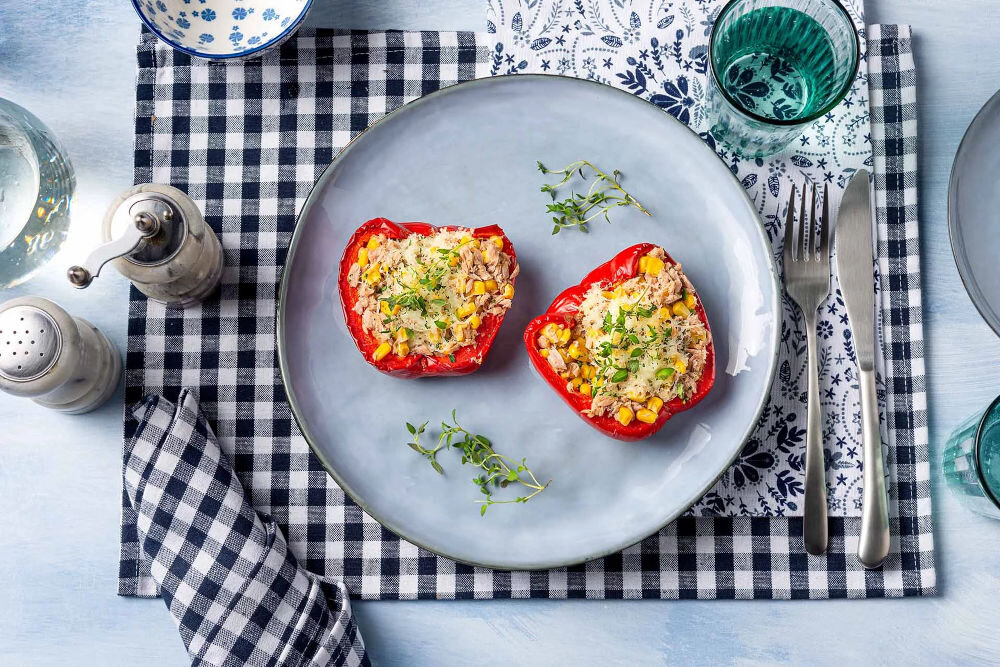 Plan dietetyczny BEDIET 3 miesiące Dwie połówki czerwonej papryki faszerowane tuńczykiem, kukurydzą i serem, podane na błękitnym talerzu na obrusie w czarno-białą kratkę.