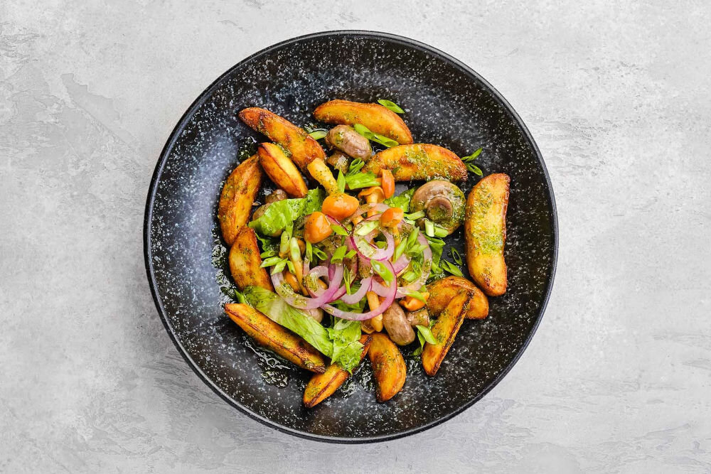 Plan dietetyczny BEDIET 12 miesięcy Pieczone cząstki ziemniaków z grzybami, czerwoną cebulą i szczypiorkiem, polane zielonym sosem ziołowym w ciemnej, nakrapianej misce.