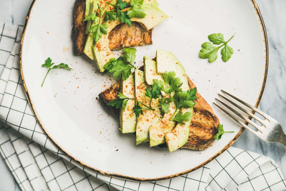 Plan dietetyczny BEDIET 12 miesięcy Dwie kromki chrupiącego tostowanego chleba z grubymi plastrami awokado, posypane papryką i udekorowane świeżą kolendrą na białym talerzu.