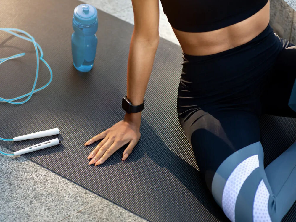 Pakiet fitness BEACTIVE 1 miesiąc Kobieta siedząca na macie do ćwiczeń obok skakanki i bidonu z wodą, ubrana w czarne legginsy i top.