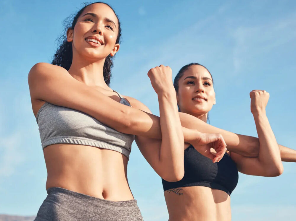 Pakiet fitness BEACTIVE 1 miesiąc Dwie uśmiechnięte kobiety wykonujące ćwiczenia rozciągające ramiona na świeżym powietrzu pod bezchmurnym niebem.