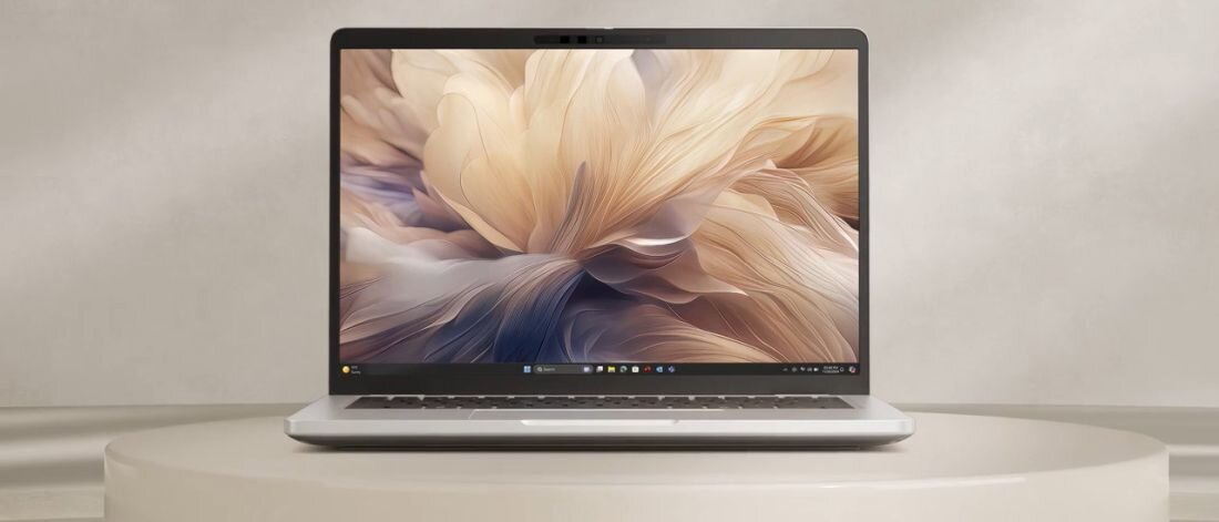 Laptop z wyświetlaczem pokazującym abstrakcyjną grafikę w odcieniach beżu i brązu, ustawiony na okrągłym stole. Na ekranie widoczny jest pasek zadań systemu Windows.