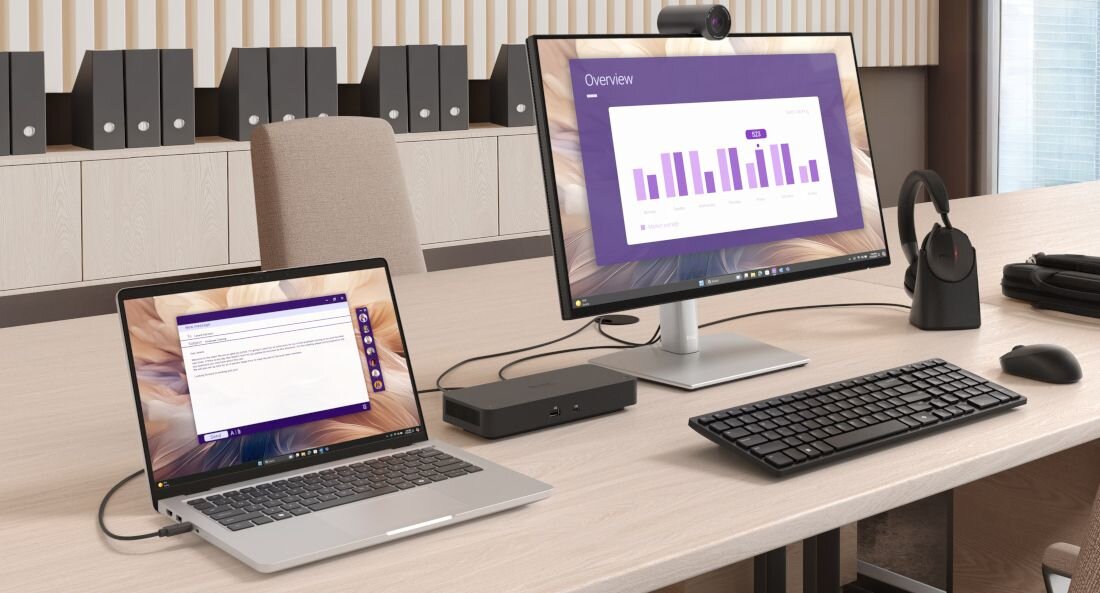 Na biurku znajduje się otwarty laptop oraz monitor z wykresem słupkowym i napisem \'Overview\'. Obok monitora leży klawiatura, myszka i stojak na słuchawki.