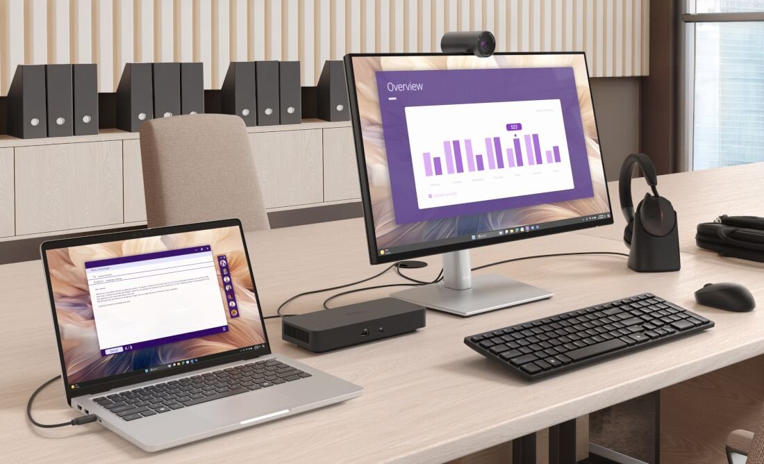 Na biurku znajduje się laptop i monitor, oba wyświetlają interfejsy z wykresami i tekstem. Obok nich leżą klawiatura, mysz i słuchawki na stojaku.