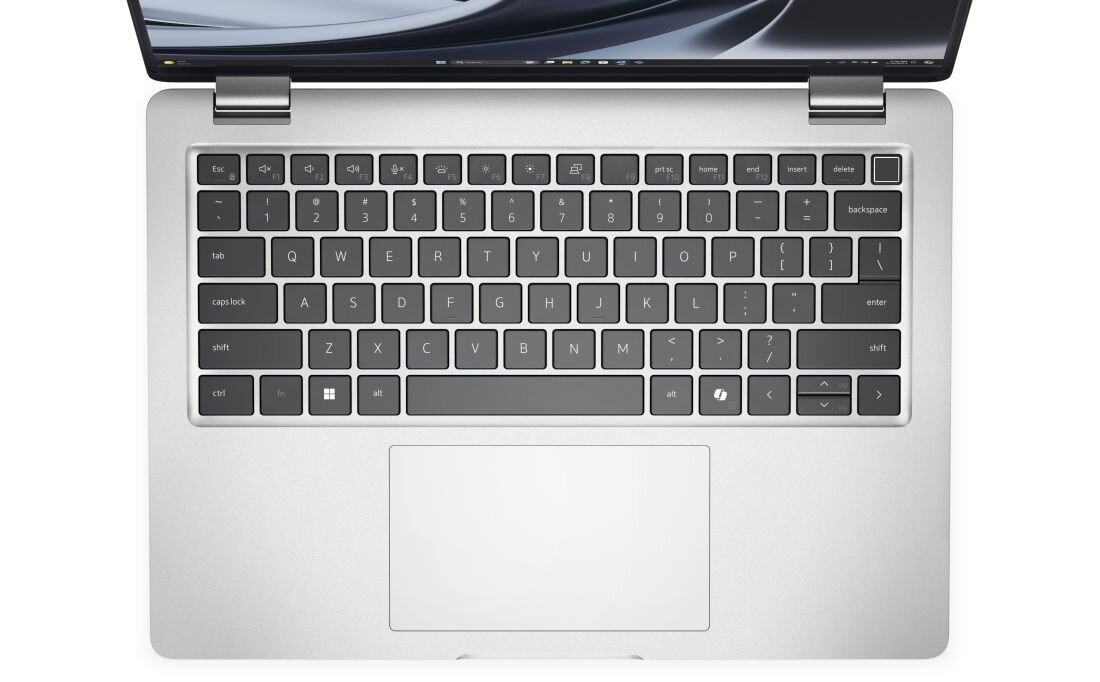 Obraz przedstawia klawiaturę laptopa z dużym touchpadem poniżej. Klawisze są ciemne z jasnymi oznaczeniami, a obudowa jest srebrna.
