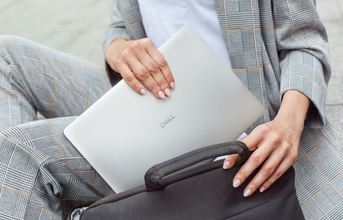 Osoba trzyma srebrny laptop z widocznym logo 'Dell', wkładając go do czarnej torby. Ubrana jest w szary garnitur w kratę.