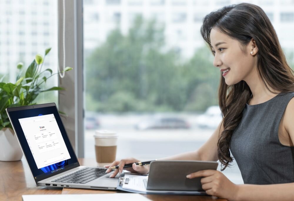 Osoba siedzi przy biurku z otwartym laptopem, na którym widoczny jest ekran z tekstem \'Hello\'. Obok laptopa znajduje się kubek z napojem i roślina w doniczce.