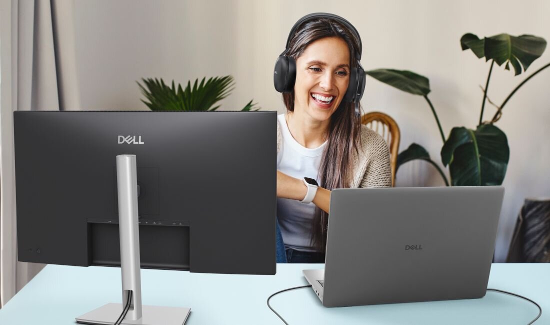 Osoba siedzi przy biurku z laptopem marki Dell i dużym monitorem tej samej marki. Na monitorze widoczne jest logo \'Dell\'.