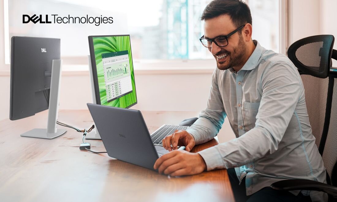 Mężczyzna siedzi przy biurku, używając laptopa marki Dell. W tle widoczny jest monitor z wykresami oraz logo \'Dell Technologies\'.