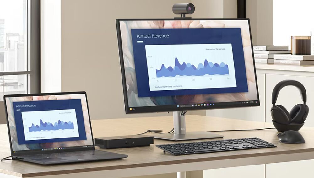 Na biurku znajduje się laptop i monitor, oba wyświetlają wykres z napisem \'Annual Revenue\'. Obok monitora leżą słuchawki, klawiatura i myszka.