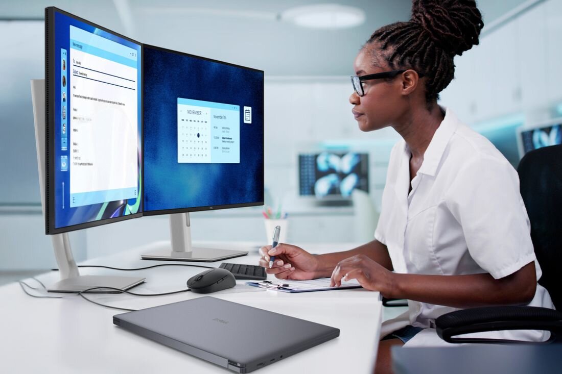 Osoba siedzi przy biurku w nowoczesnym biurze, pracując na dwóch monitorach z wyświetlonymi aplikacjami. Na biurku znajduje się zamknięty laptop z widocznym logo producenta, myszka i notatnik.