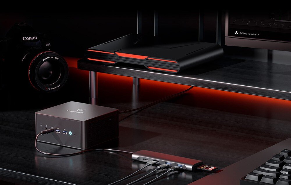 Komputer MINISFORUM UM890 Pro Mini PC podłączony do huba USB na ciemnym biurku z profesjonalnym aparatem fotograficznym w tle.
