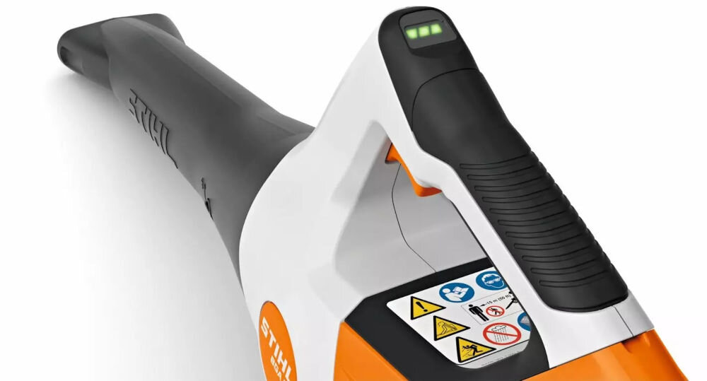 Na zdjęciu widoczna jest dmuchawa do liści z logo STIHL, z czarno-białym uchwytem i pomarańczowym korpusem. Na urządzeniu znajdują się różne symbole ostrzegawcze oraz wskaźnik naładowania baterii.