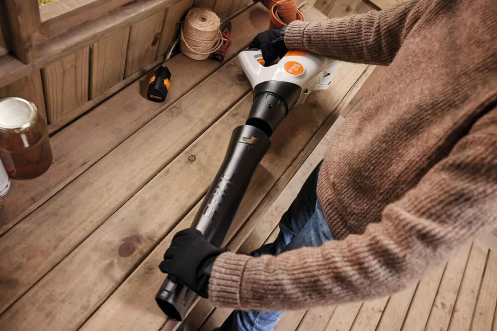 Osoba trzyma akumulatorową dmuchawę do liści z widocznym logo STIHL na drewnianej podłodze. W tle znajdują się sznurki i słoik na półce.