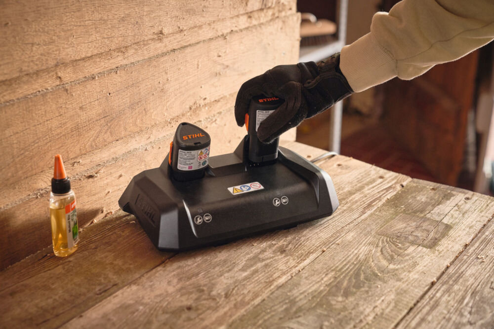Na drewnianym stole znajduje się szybka ładowarka z dwoma akumulatorami marki STIHL. Obok ładowarki stoi mała butelka z pomarańczową zakrętką.