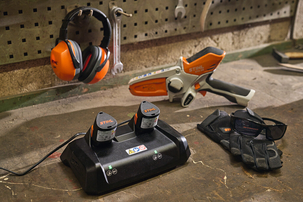 Na stole warsztatowym znajduje się szybka ładowarka z dwoma akumulatorami marki STIHL. Obok leżą rękawice ochronne, okulary oraz narzędzie ogrodnicze, a na ścianie wisi para ochronników słuchu.
