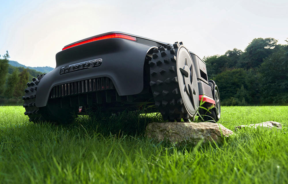 Robot koszący DREAME A3 AWD Pro 2500 trawnik robot widoczny od tyłu najeżdża kołem na kamień napęd na wszystkie koła cztery silniki w kołach może pokonywać nachylenia do 80% (38.6°) i przeszkody do 5,5 cm wysokości dwa koła mecanum dwa terenowe zderzak wpływa na bezpieczeństwo i amortyzuje ewentualne uderzenia
