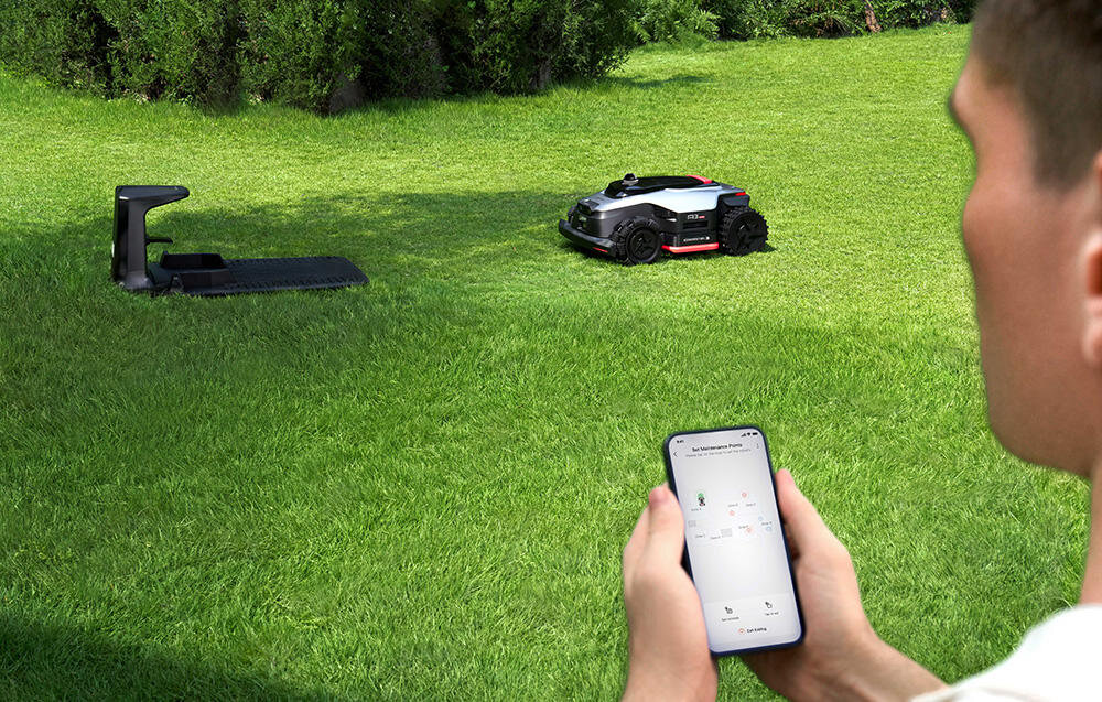 Robot koszący DREAME A3 AWD Pro 2500 trawnik robot jedzie do stacji ładowania na pierwszym planie osoba z telefonem w dłoni wysokość koszenia można dostosować w aplikacji Dreamehome w zakresie od 3 do 10 cm radzi sobie również z gęstą trawą o wysokości nawet 20 cm zmiana ustawień odbywa się błyskawicznie za pomocą jednego kliknięcia szerokość robocza wynosi 40 cm
