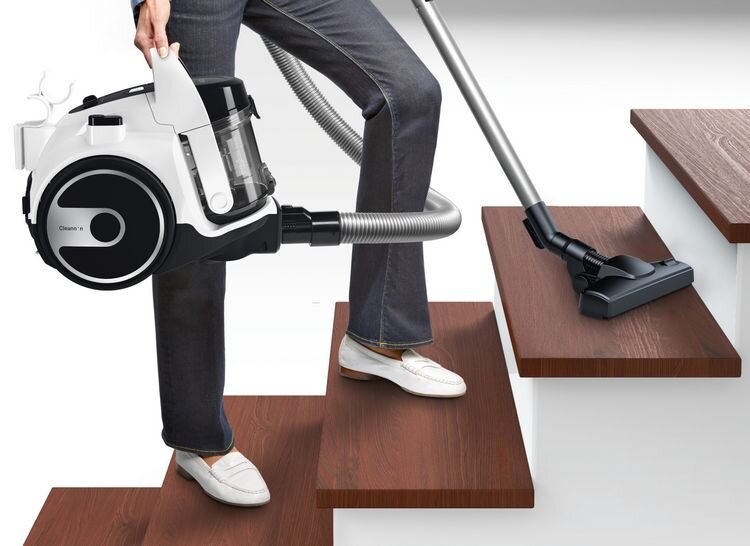 Osoba trzyma odkurzacz z przezroczystym pojemnikiem na kurz, stojąc na drewnianych schodach. Odkurzacz ma napis \'Clean\'n\' na boku i jest podłączony do metalowej rury z końcówką ssącą na schodach.