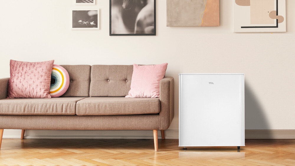 LODÓWKA TCL RF045DWE0 Na fotografii w salonie stoi biała chłodziarka TCL obok beżowej sofy z różowymi poduszkami i okrągłą, kolorową poduszką w kształcie tęczowego kręgu. Nad sofą zawieszono kilka obrazów i grafik w ramkach, a podłogę pokrywa parkiet w jodełkę