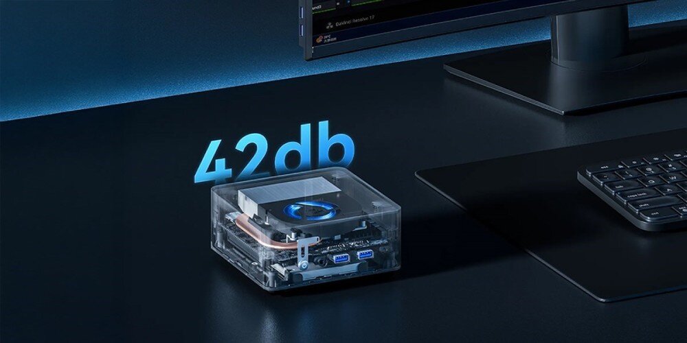 Komputer QOOBE II AP13620 Przezroczysty mini PC z widocznym chłodzeniem i podświetleniem, na biurku obok klawiatury z niebiesym napisem '42 db' nad urządzeniem, porty, gniazda, łączność