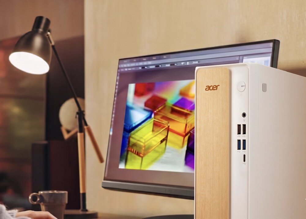 Na biurku znajduje się komputer stacjonarny z logo \'acer\' na przednim panelu oraz monitor wyświetlający kolorową grafikę. Obok komputera stoi lampa biurkowa i kubek.
