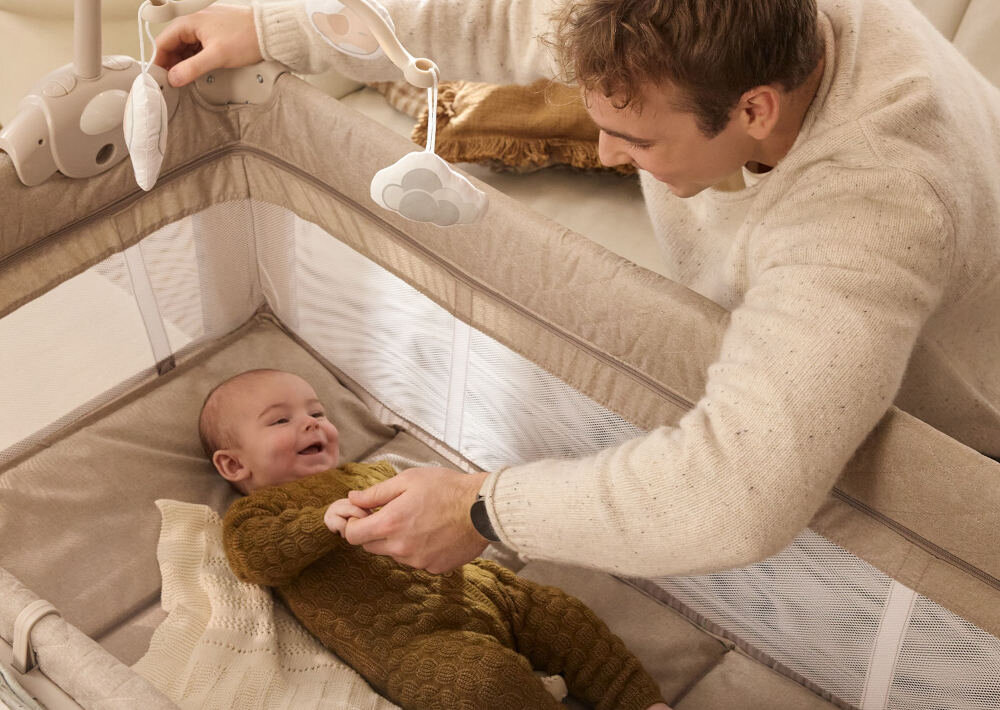 Łóżeczko turystyczne KINDERKRAFT Leody Up Beżowy Mobilne wymiary wysokość 79 cm, szerokość 22,5 cm ma wygodną przestrzeń do snu i zabawy osoba nachyla się nad dzieckiem w łóżeczku dziecko w łóżeczku się uśmiecha