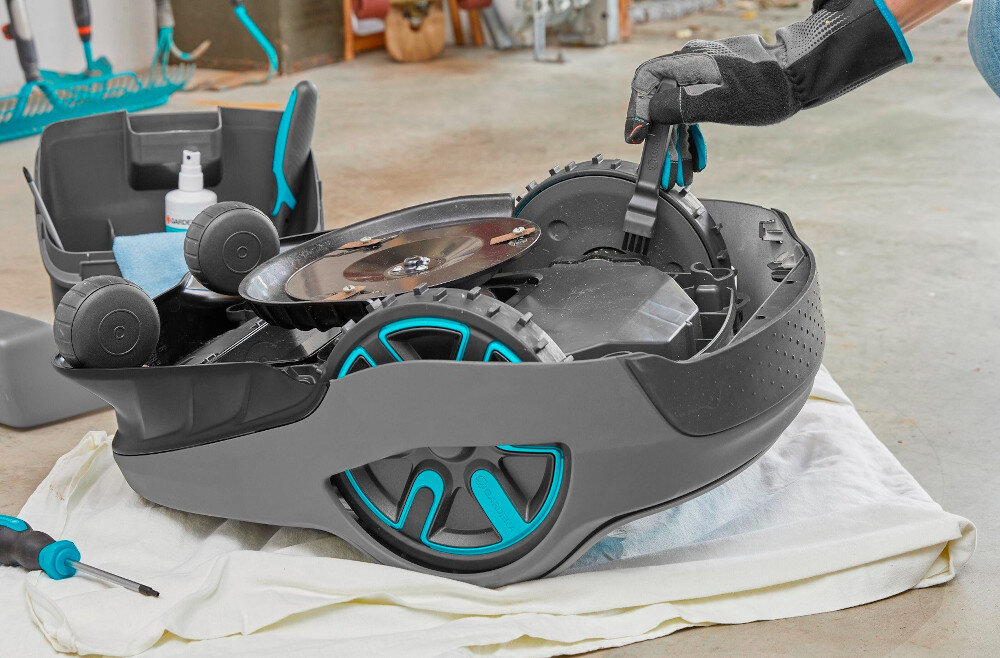 Zestaw do konserwacji GARDENA robotów koszących 4067-20 garaż podłoga robot leży na podłodze osoba czyści koła za pomocą małej szczoteczki mała szczotka do precyzyjnego usuwania drobnych zabrudzeń wkrętak umożliwiający samodzielną wymianę ostrzy pełna kontrola nad stanem technicznym robota