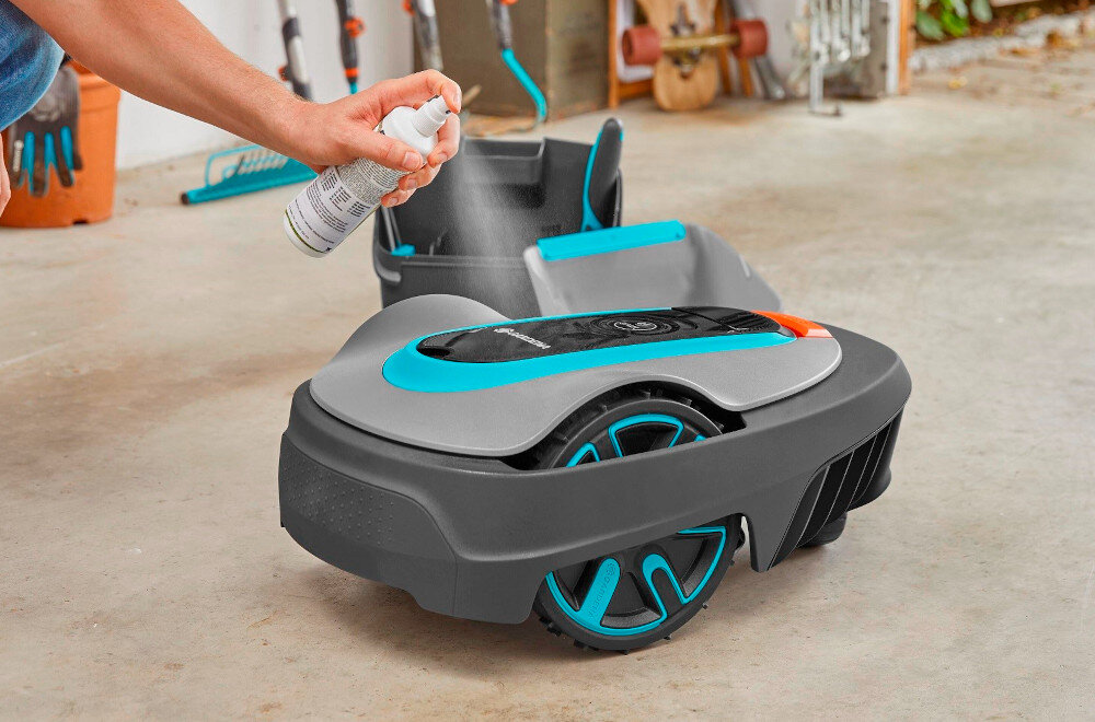 Zestaw do konserwacji GARDENA robotów koszących 4067-20 garaż podłoga osoba spryskuje sprayem robota koszącego w tle narzędzia wiadro spray do pielęgnacji tworzywa sztucznego ochrona przed starzeniem i matowieniem ułatwia utrzymanie estetycznego wyglądu urządzenia minimalizuje ryzyko uszkodzeń mechanicznych