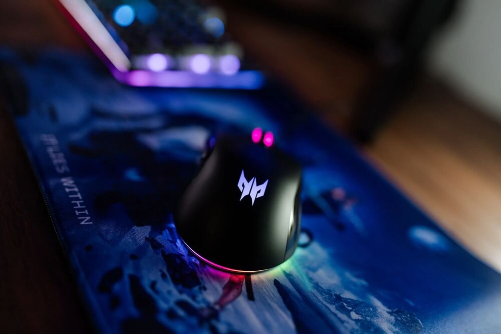Na obrazie widoczna jest czarna mysz komputerowa z podświetleniem RGB i logo PREDATOR na podkładce z grafiką. W tle znajduje się klawiatura z podświetleniem.