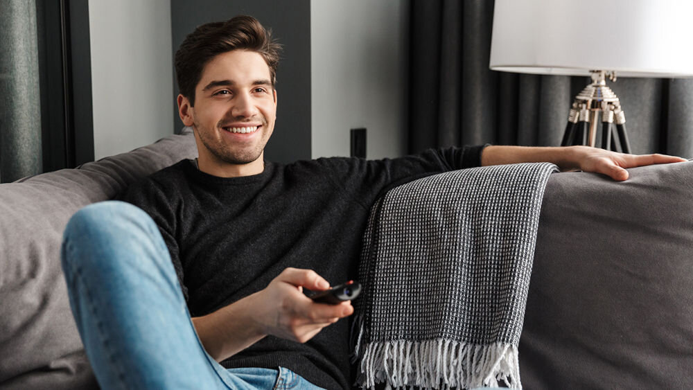 Soundbar SHARP HT-SBW55121 HDMI eARC/CEC korektor tryby nocne regulacja dźwięku, osoba siedząca na kanapie z pilotem w ręku