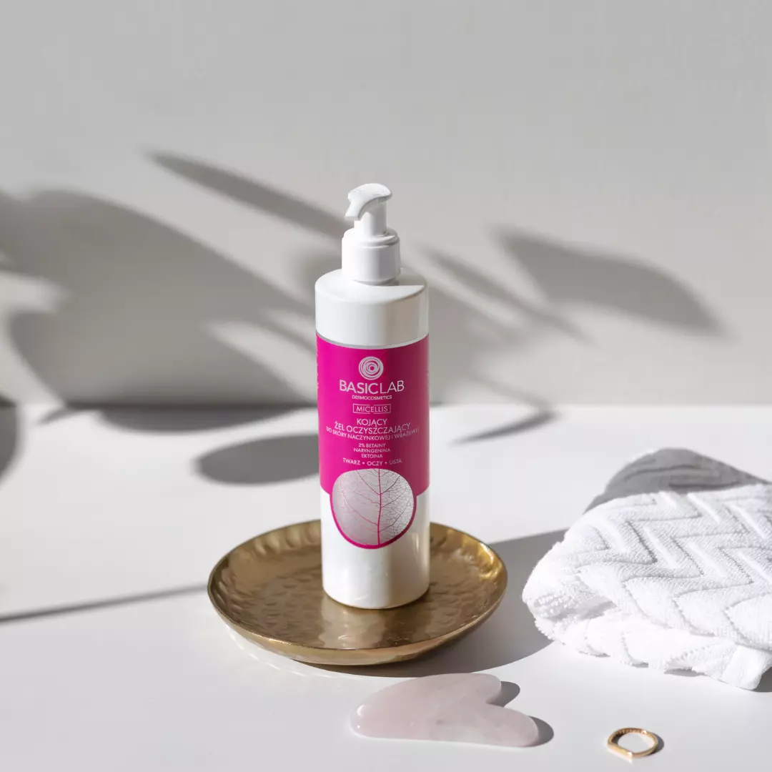 Biała butelka z pompką, na której widnieje różowa etykieta z napisem \'BASICLAB DERMOCOSMETICS KOJĄCY ŻEL OCZYSZCZAJĄCY\'. Obok znajduje się złota tacka, biały ręcznik i różowy kamień w kształcie serca.