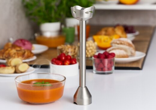 Przystawka miksująca na kuchennym blacie. Obok niej miska z zupą. W tle różne przekąski, owoce i warzywa w miseczkach.