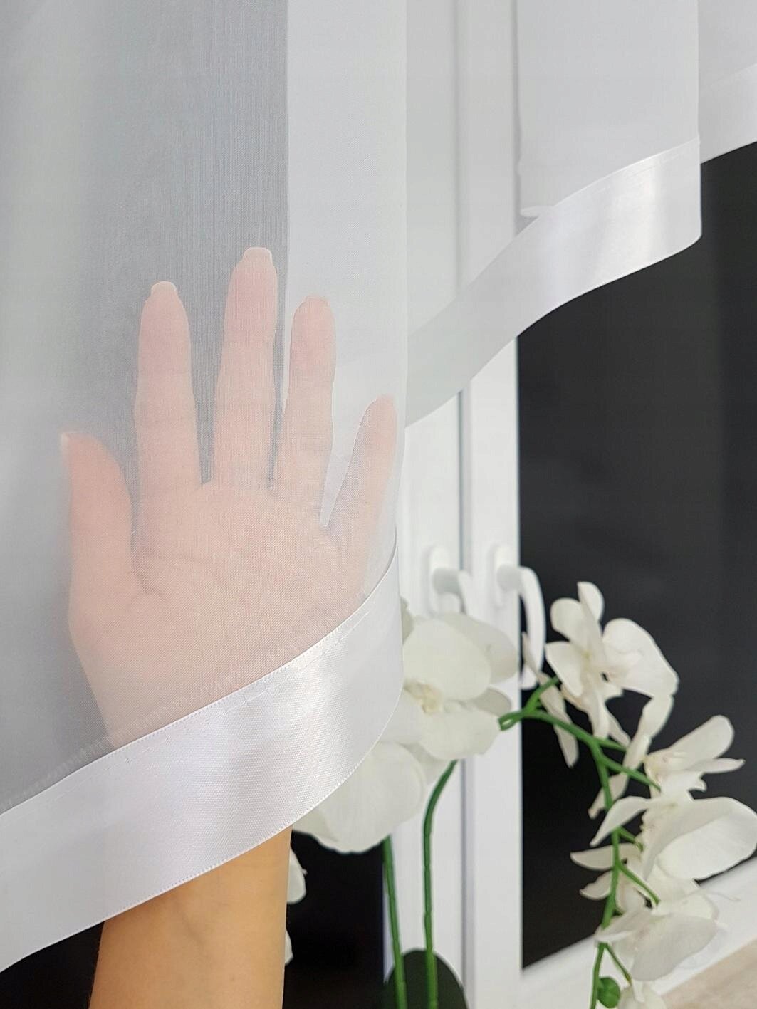 Na zdjęciu widać rękę dotykającą przezroczystej, białej firany z woalu z satynowym wykończeniem. W tle znajduje się okno oraz białe kwiaty.