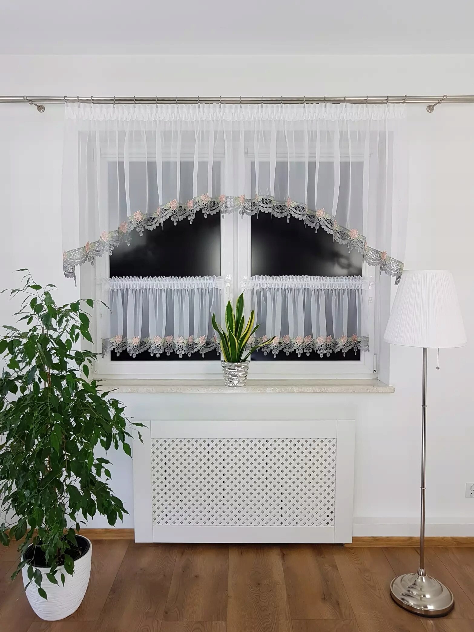 Na zdjęciu widoczne jest okno z białymi firanami zdobionymi szarą gipiurą. Przed oknem stoi roślina doniczkowa, a obok znajduje się lampa stojąca.