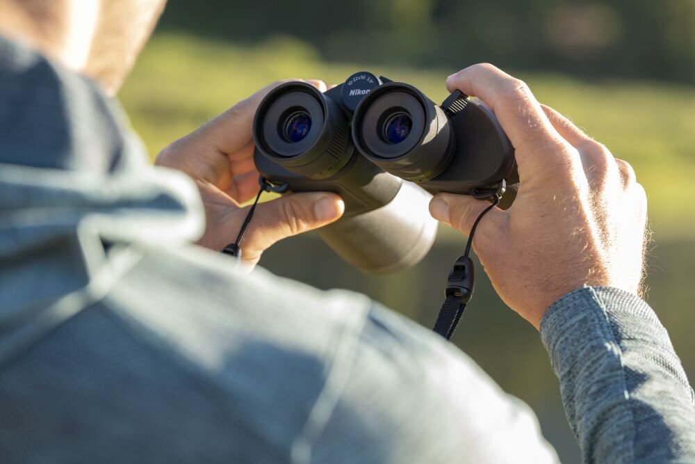 Osoba trzyma lornetkę marki Nikon, skierowaną w stronę otoczenia na zewnątrz. Widoczne są dłonie oraz fragment odzieży wierzchniej.