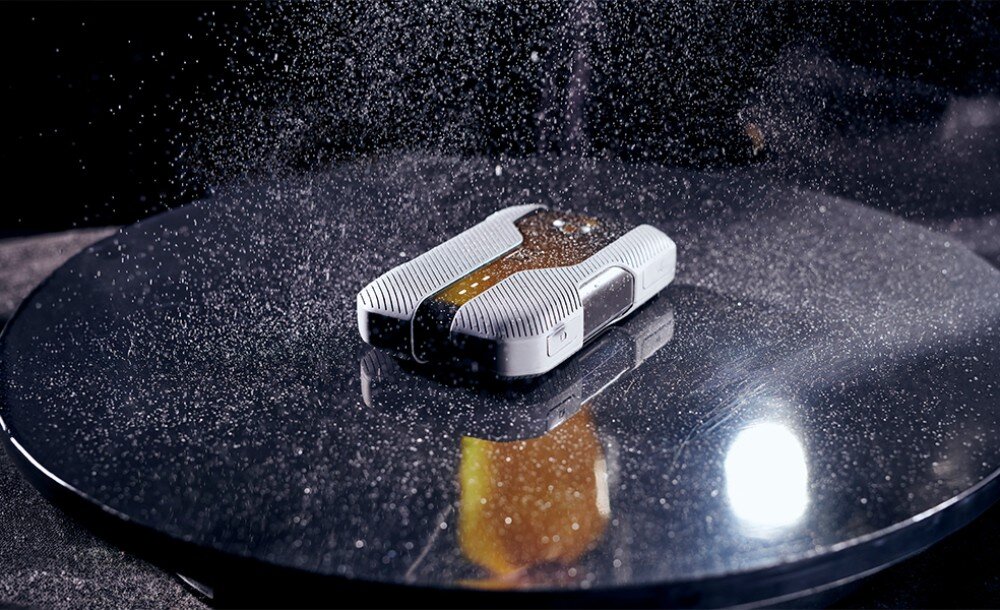 Router CAT CQ10-SAB-RON-NN 5G Widok na jasne urządzenie o wzmocnionej obudowie, które spoczywa na czarnej, błyszczącej powierzchni pod strumieniem drobnych kropel wody