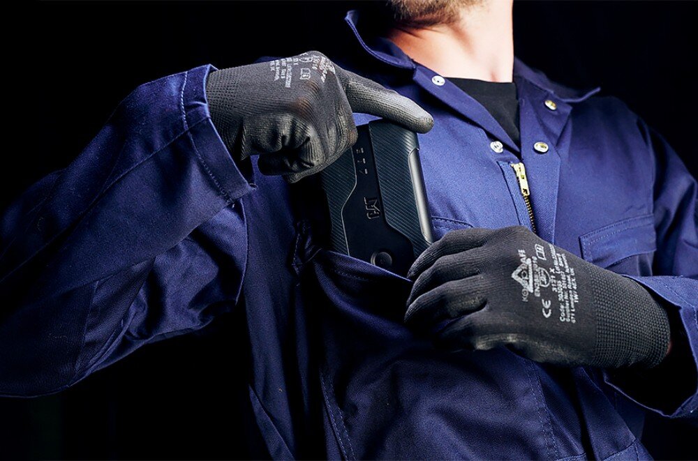 Router CAT CQ10-SAB-RON-NN 5G Osoba w niebieskim ubraniu roboczym i ciemnych rękawicach ochronnych chowa płaskie urządzenie do kieszeni na piersi