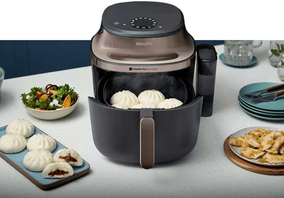 Na blacie kuchennym stoi frytkownica beztłuszczowa marki PHILIPS z otwartą szufladą, w której znajdują się bułeczki. Obok urządzenia widoczne są talerze z sałatką i pierożkami oraz stos talerzy z widelcami.