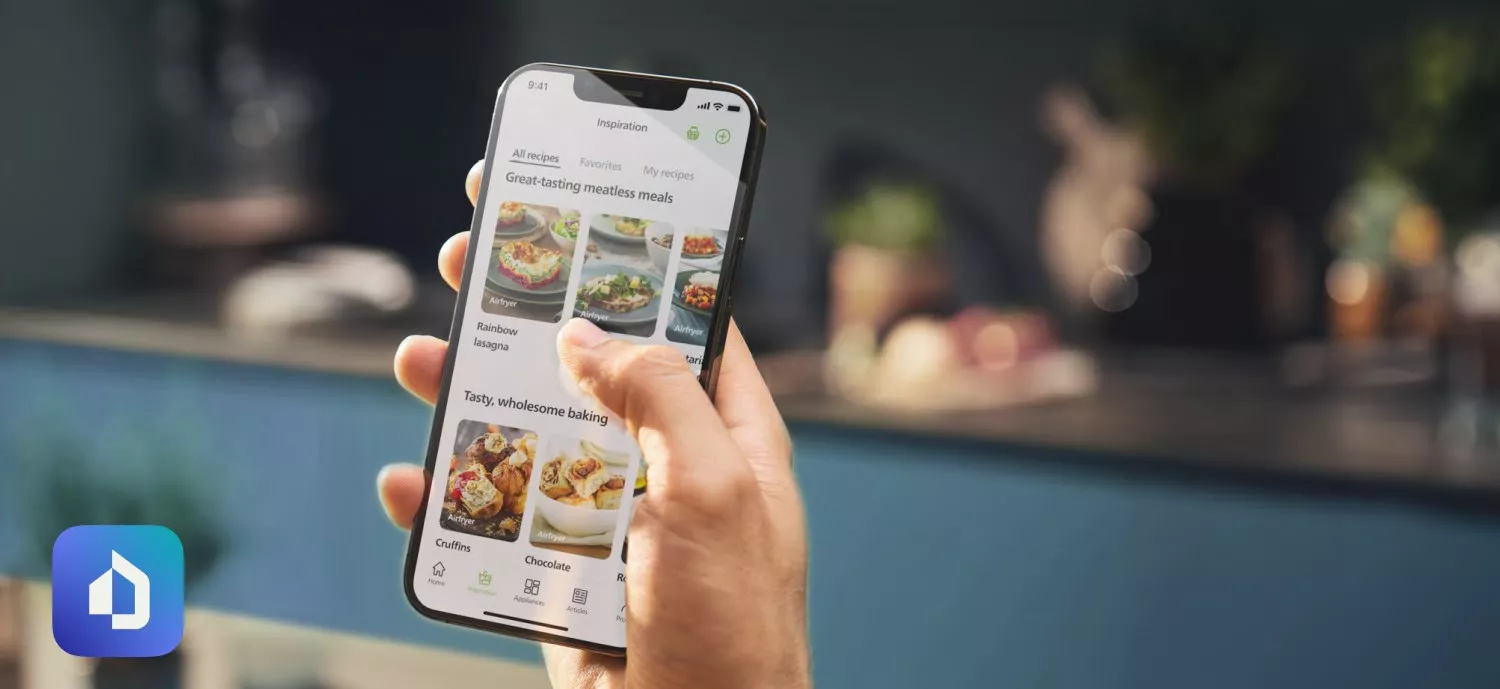 Osoba trzyma smartfon z aplikacją wyświetlającą przepisy kulinarne, w tym \'Great-tasting meatless meals\' i \'Tasty, wholesome baking\'. W tle widoczna jest rozmyta kuchnia.