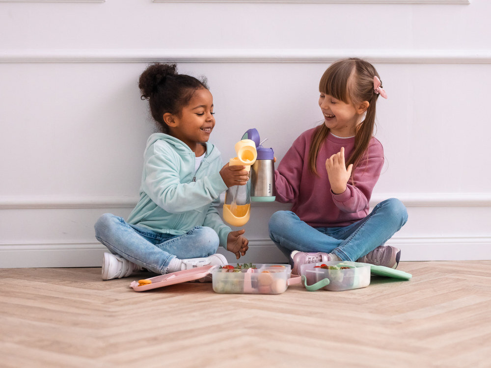 Dwoje dzieci siedzi na podłodze, trzymając kolorowe butelki i pojemniki na jedzenie. Dzieci są uśmiechnięte i wyglądają na zadowolone.