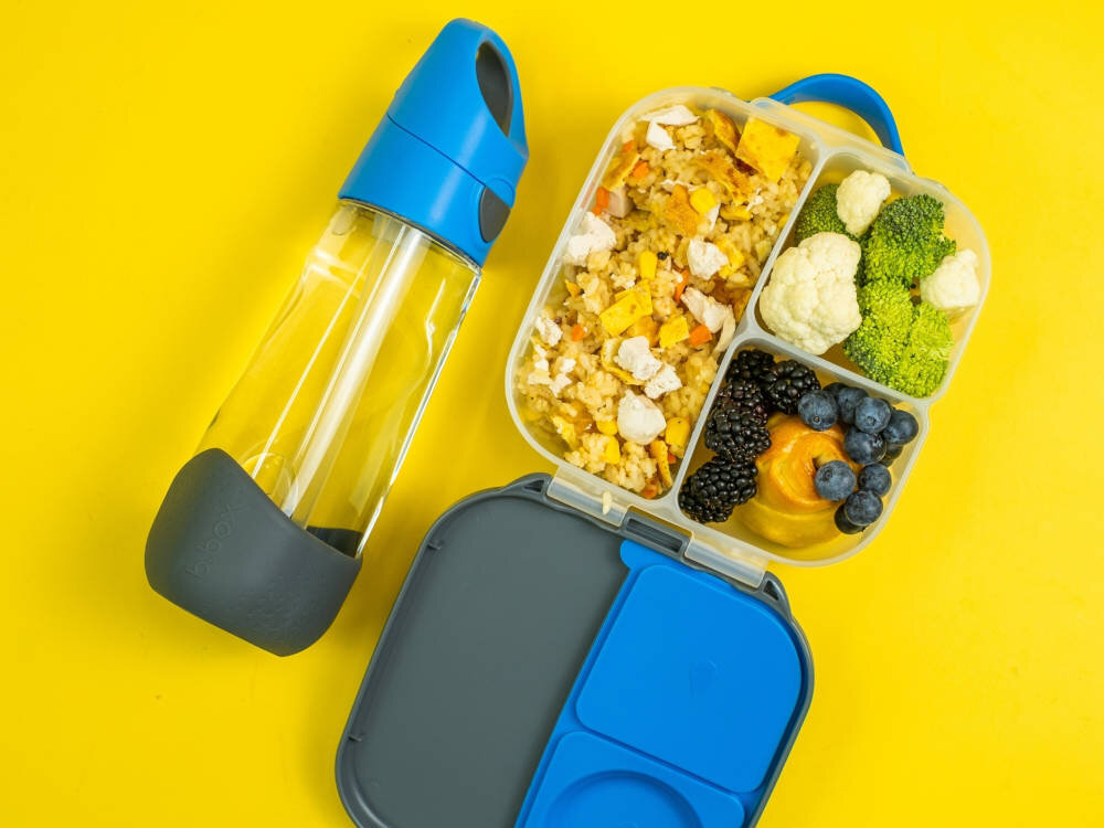 Na żółtym tle znajduje się przezroczysta butelka z niebieskim korkiem i szarym uchwytem oraz otwarty pojemnik na żywność z różnymi produktami spożywczymi. W pojemniku widoczne są kawałki kurczaka z ryżem, brokuły, kalafior, jeżyny, borówki i kawałki owoców.