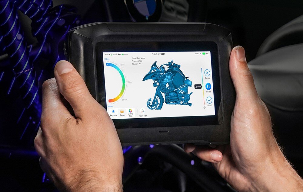 Osoba trzyma urządzenie z ekranem dotykowym, na którym wyświetlany jest model 3D motocykla oraz wykresy i opcje menu. W tle widoczne są elementy wnętrza pojazdu.
